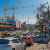 Stau auf einer mehrspurigen Straße mit Baustelle und Kränen im Hintergrund. Verkehrsschilder weisen auf Rathaus und Dorothy Quartier hin. Ein Roller parkt am Straßenrand.