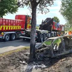 Feuerwehr im Einsatz nach einem **Unfall mit einem Elektroauto**. Ein ausgebranntes Fahrzeug liegt neben der Straße, ein LKW der Feuerwehr steht bereit.