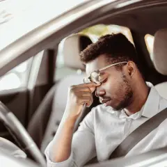 Erschöpfter Mann mit Brille reibt sich die Augen, während er im Auto sitzt. Lange Fahrt, tavor autofahren.