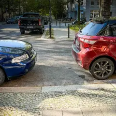 Zwei Autos parken nebeneinander, ein blauer Audi und ein roter Kleinwagen, am abgesenkten Bordstein.