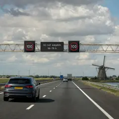 Autobahn in den Niederlanden mit einem Blitzer-Schild, das 100 km/h bei nasser Fahrbahn zwischen 16-19 Uhr anzeigt. Ein Windrad und Autos sind zu sehen.