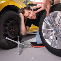Mann beim Reifenwechsel, der frustriert nachdenkt, ob die Handbremse angezogen ist. Ein Rad ist ab, das andere liegt daneben.