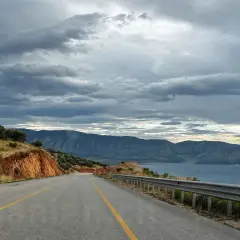Eine Straße führt durch eine bergige Landschaft mit Olivenbäumen und Blick auf das Meer. Die Wolken sind dramatisch. Maut Albanien.