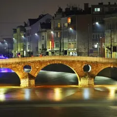 Eine beleuchtete Brücke in Sarajevo, Bosnien, bei Nacht. Die Lichter der Stadt spiegeln sich im Wasser.