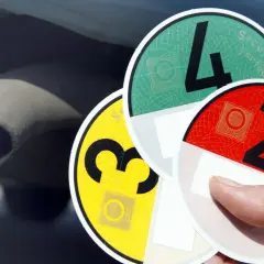 Hand hält Umweltplaketten: eine gelbe Plakette mit der Zahl 3, eine grüne mit der 4 und eine rote mit der 2. Im Hintergrund ist ein Auspuff zu sehen.