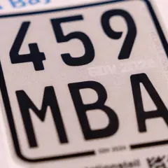 Nahaufnahme eines silbernen Nummernschilds mit schwarzer Schrift: 459 MBA. Ein Teil eines blauen Logos und Text sind sichtbar.