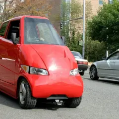 Roter, komischer Kleinwagen mit einer ungewöhnlichen Form fährt auf der Straße. Daneben ein silbernes Auto.