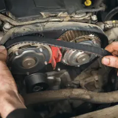 Hände montieren einen Zahnriemen an Zahnrädern im Motorraum. Der Zahnriemen synchronisiert Nockenwelle und Kurbelwelle, was macht der zahnriemen.
