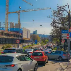 Stau auf einer mehrspurigen Straße mit Baustelle und Kränen im Hintergrund. Verkehrsschilder weisen auf Rathaus und Dorothy Quartier hin. Ein Roller parkt am Straßenrand.