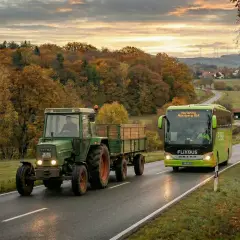 Ein grüner Traktor mit Anhänger und ein Flixbus fahren auf einer Landstraße im Herbst. Die 132 bkat Szene zeigt bunte Bäume und eine malerische Landschaft.