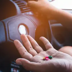 Hand hält Pillen, darunter Ibuprofen, vor einem Lenkrad. Vorsicht beim Autofahren mit Medikamenten!