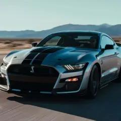 Silberner Ford Mustang Shelby GT500 mit schwarzen Streifen fährt auf einer Straße in der Wüste.