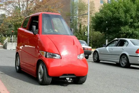 Roter, komischer Kleinwagen mit einer ungewöhnlichen Form fährt auf der Straße. Daneben ein silbernes Auto.
