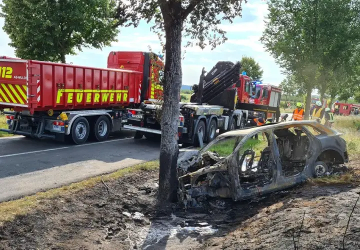 Feuerwehr im Einsatz nach einem **Unfall mit einem Elektroauto**. Ein ausgebranntes Fahrzeug liegt neben der Straße, ein LKW der Feuerwehr steht bereit.