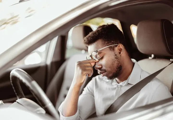 Erschöpfter Mann mit Brille reibt sich die Augen, während er im Auto sitzt. Lange Fahrt, tavor autofahren.