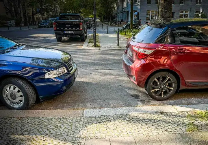 Zwei Autos parken nebeneinander, ein blauer Audi und ein roter Kleinwagen, am abgesenkten Bordstein.