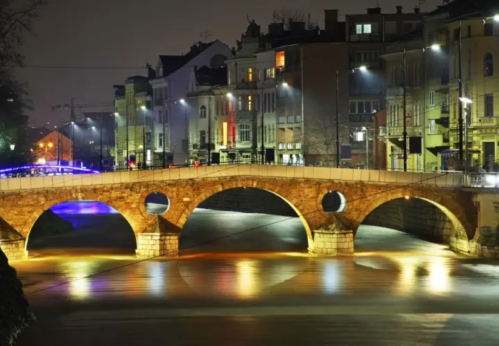 Eine beleuchtete Brücke in Sarajevo, Bosnien, bei Nacht. Die Lichter der Stadt spiegeln sich im Wasser.