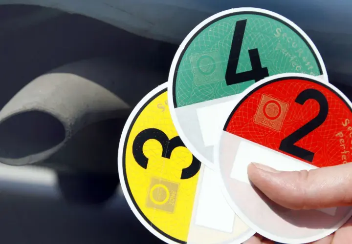 Hand hält Umweltplaketten: eine gelbe Plakette mit der Zahl 3, eine grüne mit der 4 und eine rote mit der 2. Im Hintergrund ist ein Auspuff zu sehen.