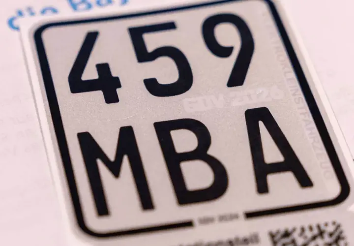 Nahaufnahme eines silbernen Nummernschilds mit schwarzer Schrift: 459 MBA. Ein Teil eines blauen Logos und Text sind sichtbar.