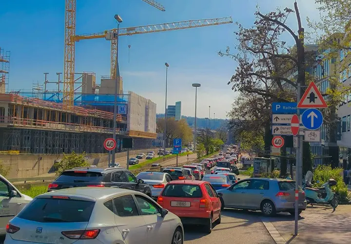 Stau auf einer mehrspurigen Straße mit Baustelle und Kränen im Hintergrund. Verkehrsschilder weisen auf Rathaus und Dorothy Quartier hin. Ein Roller parkt am Straßenrand.