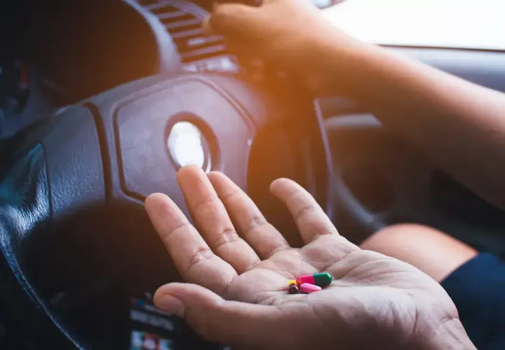Hand hält Pillen, darunter Ibuprofen, vor einem Lenkrad. Vorsicht beim Autofahren mit Medikamenten!