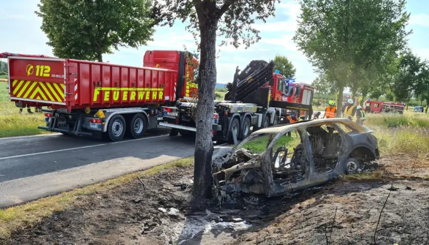 Feuerwehr im Einsatz nach einem **Unfall mit einem Elektroauto**. Ein ausgebranntes Fahrzeug liegt neben der Straße, ein LKW der Feuerwehr steht bereit.
