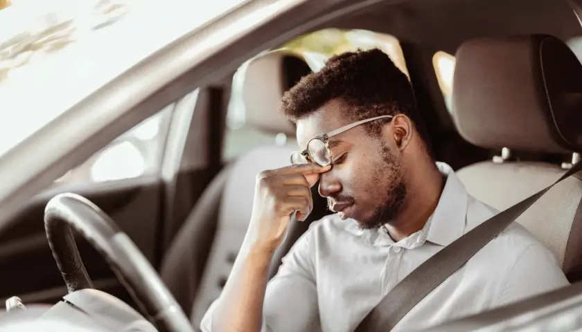 Erschöpfter Mann mit Brille reibt sich die Augen, während er im Auto sitzt. Lange Fahrt, tavor autofahren.