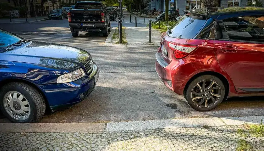 Zwei Autos parken nebeneinander, ein blauer Audi und ein roter Kleinwagen, am abgesenkten Bordstein.