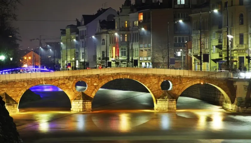 Eine beleuchtete Brücke in Sarajevo, Bosnien, bei Nacht. Die Lichter der Stadt spiegeln sich im Wasser.
