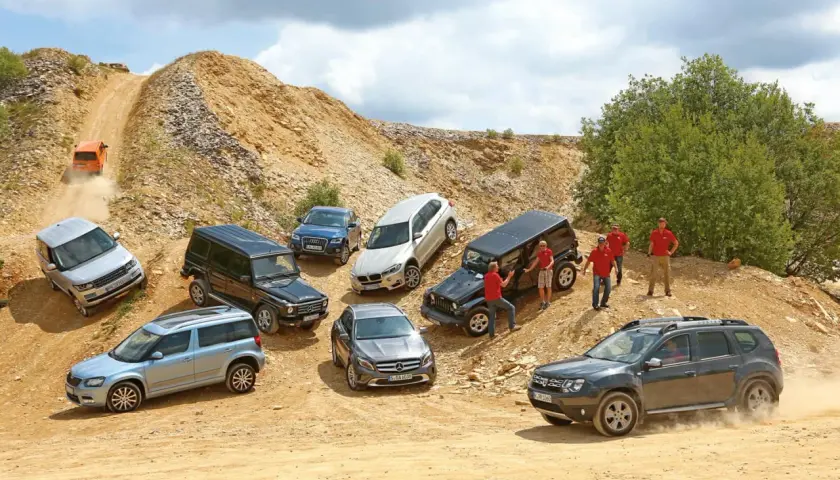 Verschiedene SUVs und Geländewagen auf einer Schotterpiste. Ein orangefarbener Wagen fährt einen steilen Hang hinauf. Das zeigt, was bedeutet offroad.