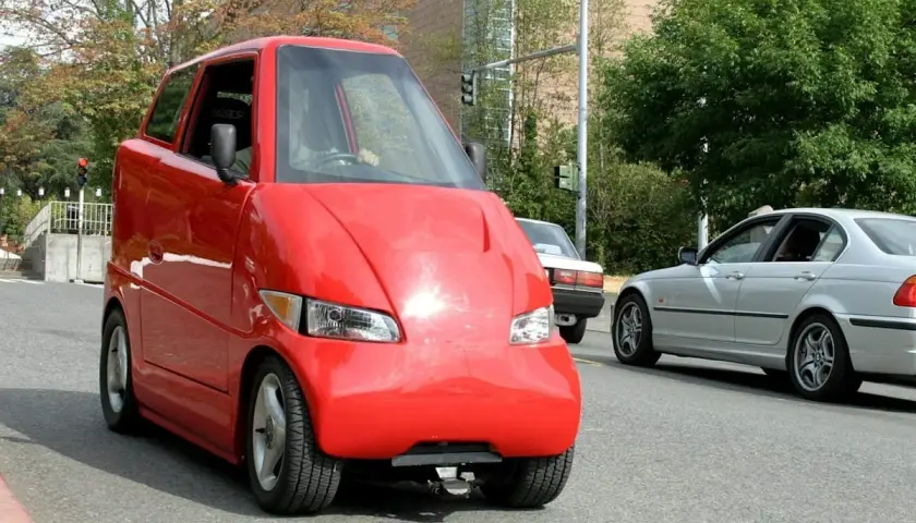 Roter, komischer Kleinwagen mit einer ungewöhnlichen Form fährt auf der Straße. Daneben ein silbernes Auto.