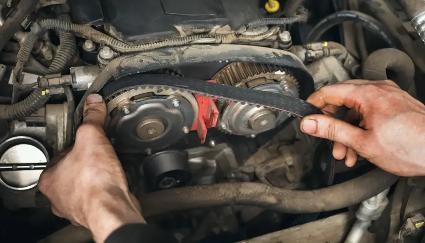 Hände montieren einen Zahnriemen an Zahnrädern im Motorraum. Der Zahnriemen synchronisiert Nockenwelle und Kurbelwelle, was macht der zahnriemen.