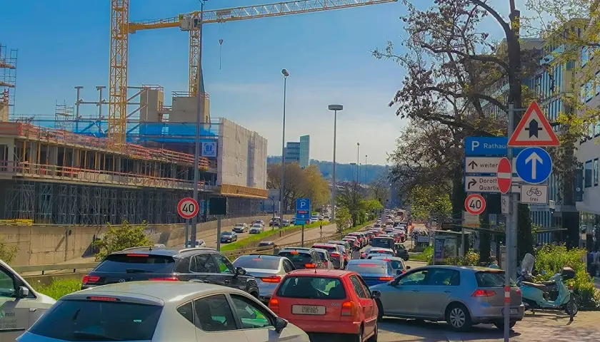 Stau auf einer mehrspurigen Straße mit Baustelle und Kränen im Hintergrund. Verkehrsschilder weisen auf Rathaus und Dorothy Quartier hin. Ein Roller parkt am Straßenrand.