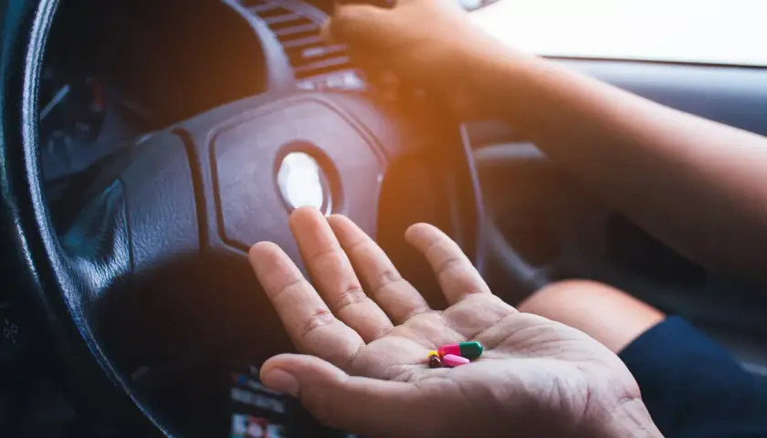 Hand hält Pillen, darunter Ibuprofen, vor einem Lenkrad. Vorsicht beim Autofahren mit Medikamenten!