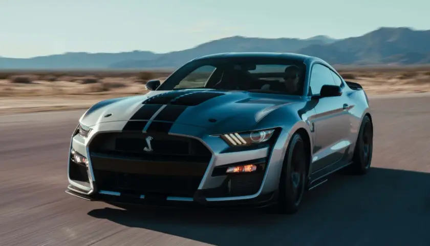 Silberner Ford Mustang Shelby GT500 mit schwarzen Streifen fährt auf einer Straße in der Wüste.