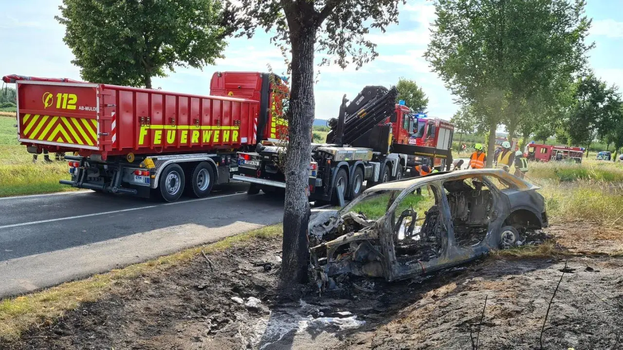 Feuerwehr im Einsatz nach einem **Unfall mit einem Elektroauto**. Ein ausgebranntes Fahrzeug liegt neben der Straße, ein LKW der Feuerwehr steht bereit.
