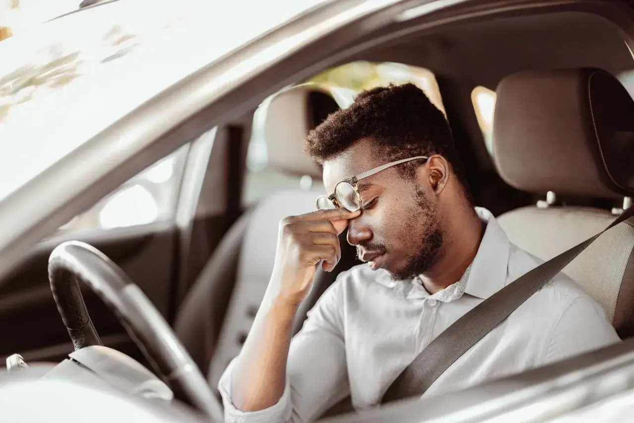 Erschöpfter Mann mit Brille reibt sich die Augen, während er im Auto sitzt. Lange Fahrt, tavor autofahren.