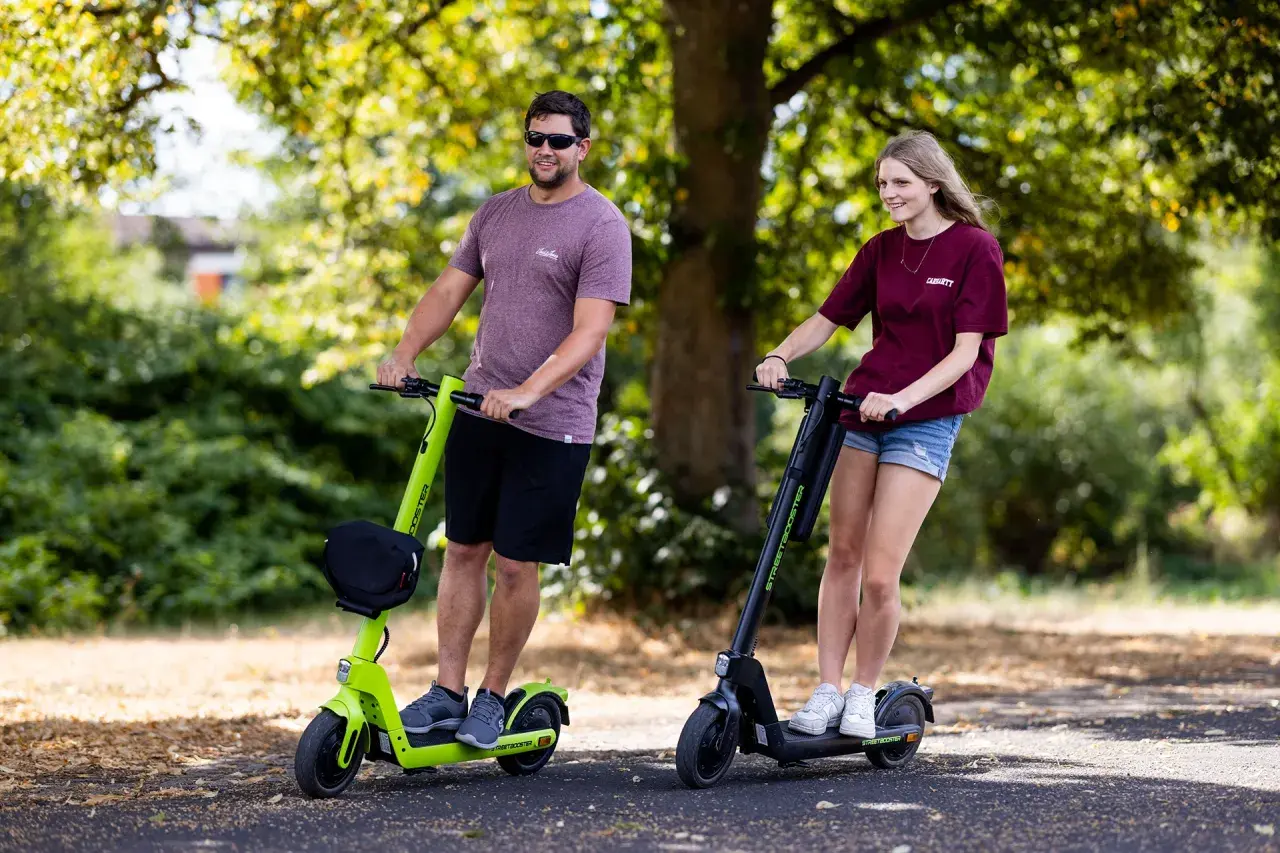 Ein Mann und eine Frau fahren nebeneinander auf E-Scootern. Sie sind im Park unterwegs, ein Ausflug mit dem e-scooter zu zweit mit kind.