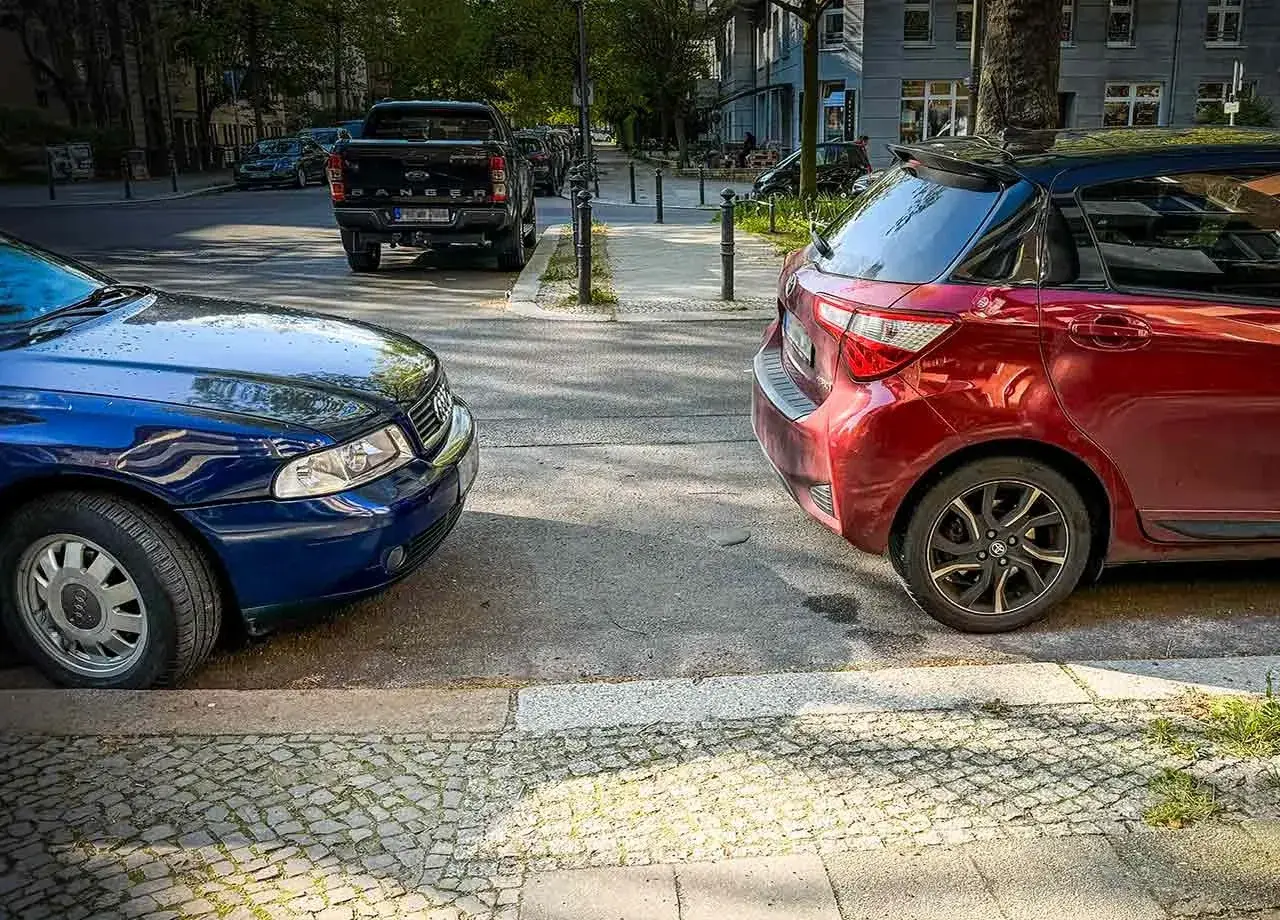 Zwei Autos parken nebeneinander, ein blauer Audi und ein roter Kleinwagen, am abgesenkten Bordstein.