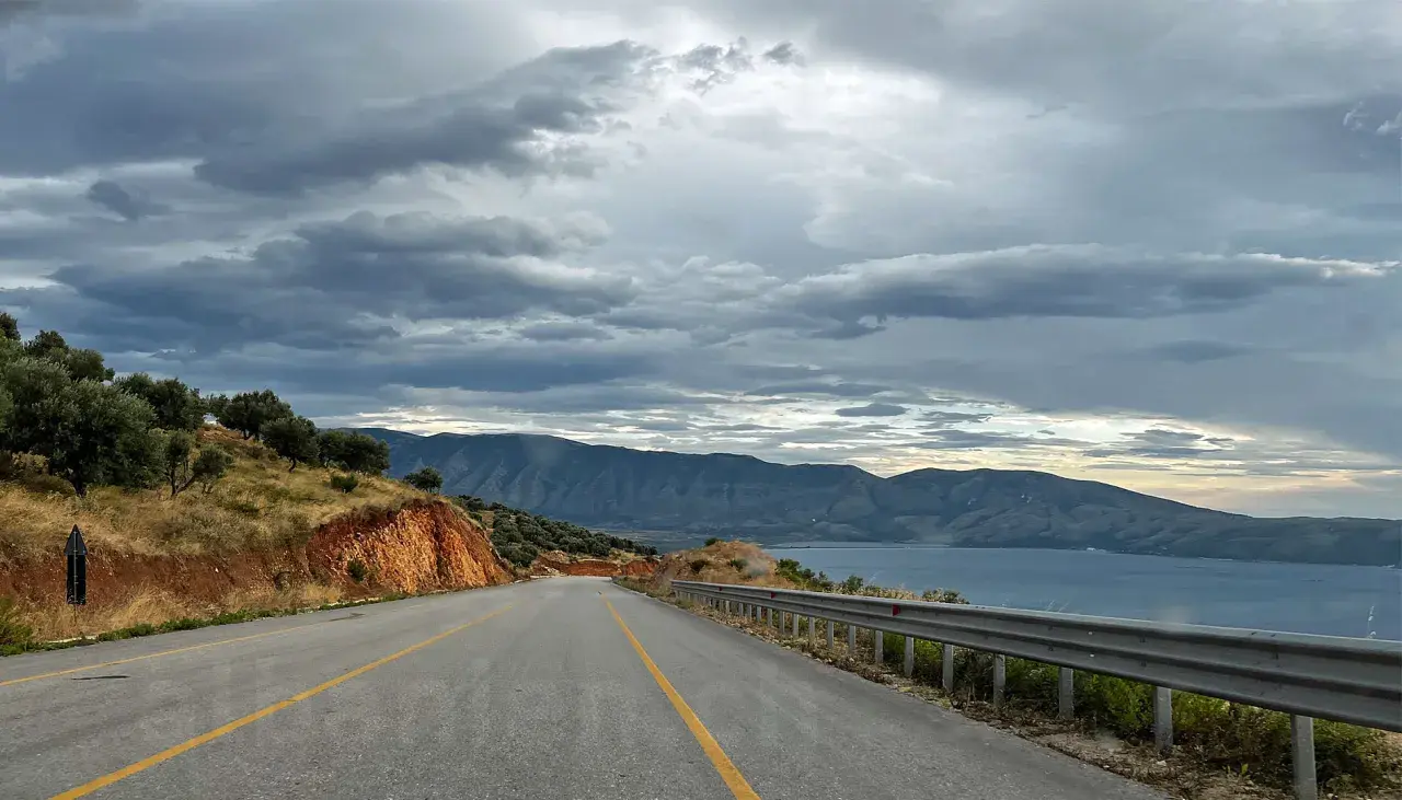Eine Straße führt durch eine bergige Landschaft mit Olivenbäumen und Blick auf das Meer. Die Wolken sind dramatisch. Maut Albanien.