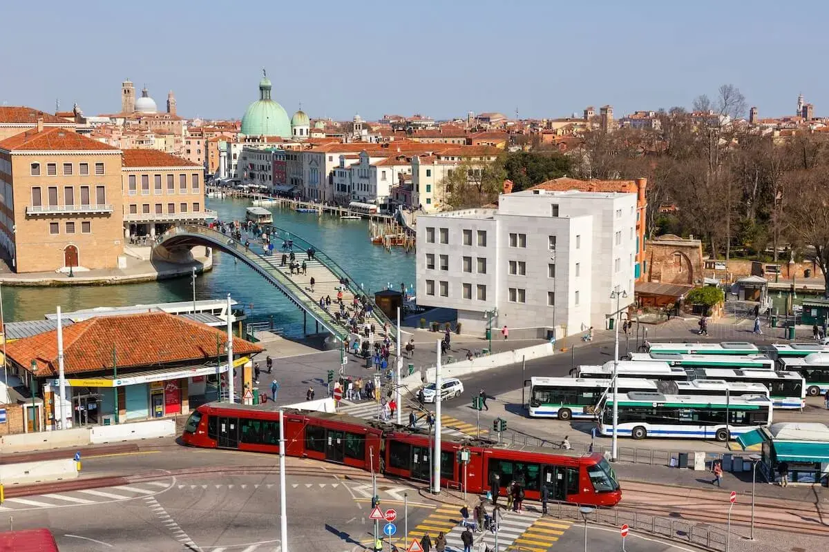 Roter Zug und Busse am Bahnhof von Venedig. Menschen überqueren eine Brücke über den Kanal.