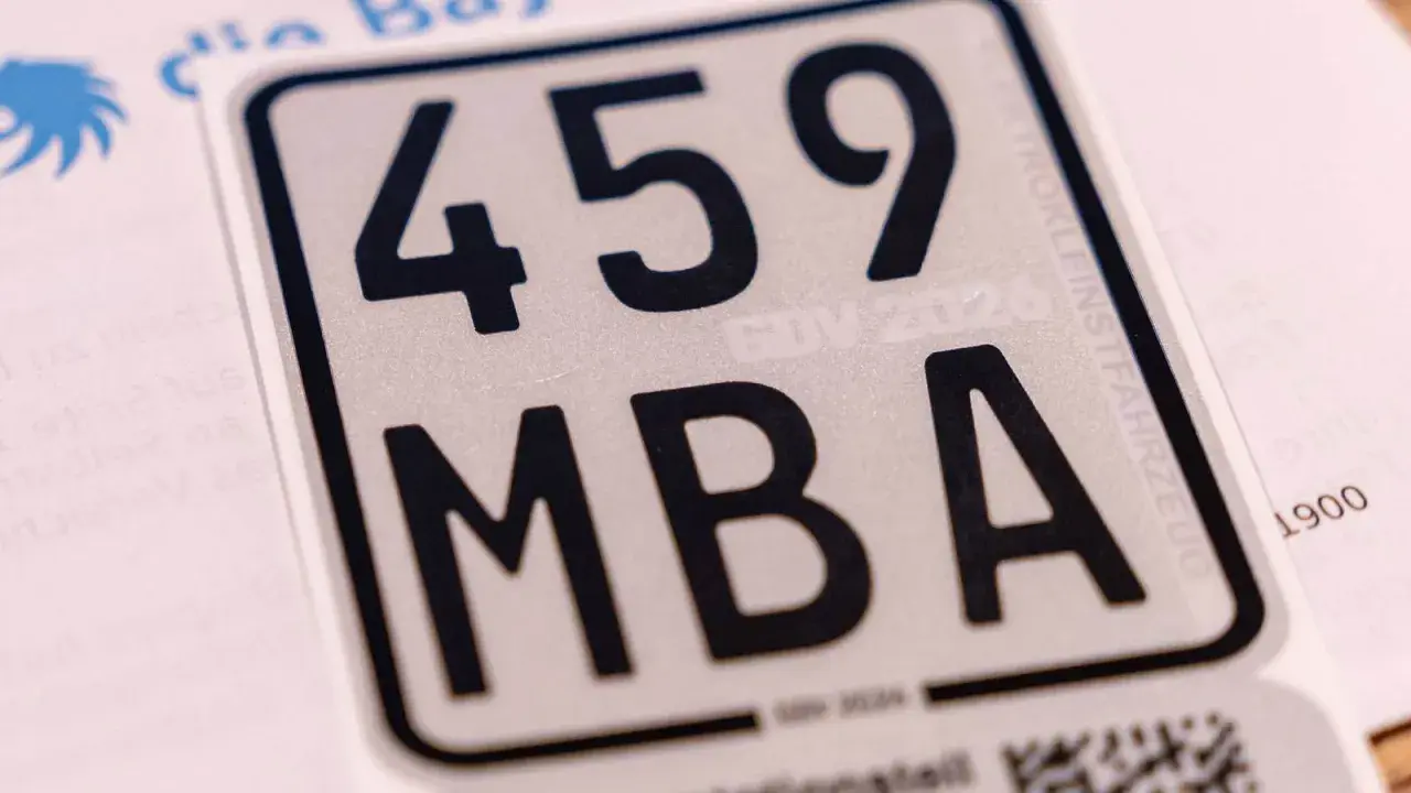Nahaufnahme eines silbernen Nummernschilds mit schwarzer Schrift: 459 MBA. Ein Teil eines blauen Logos und Text sind sichtbar.
