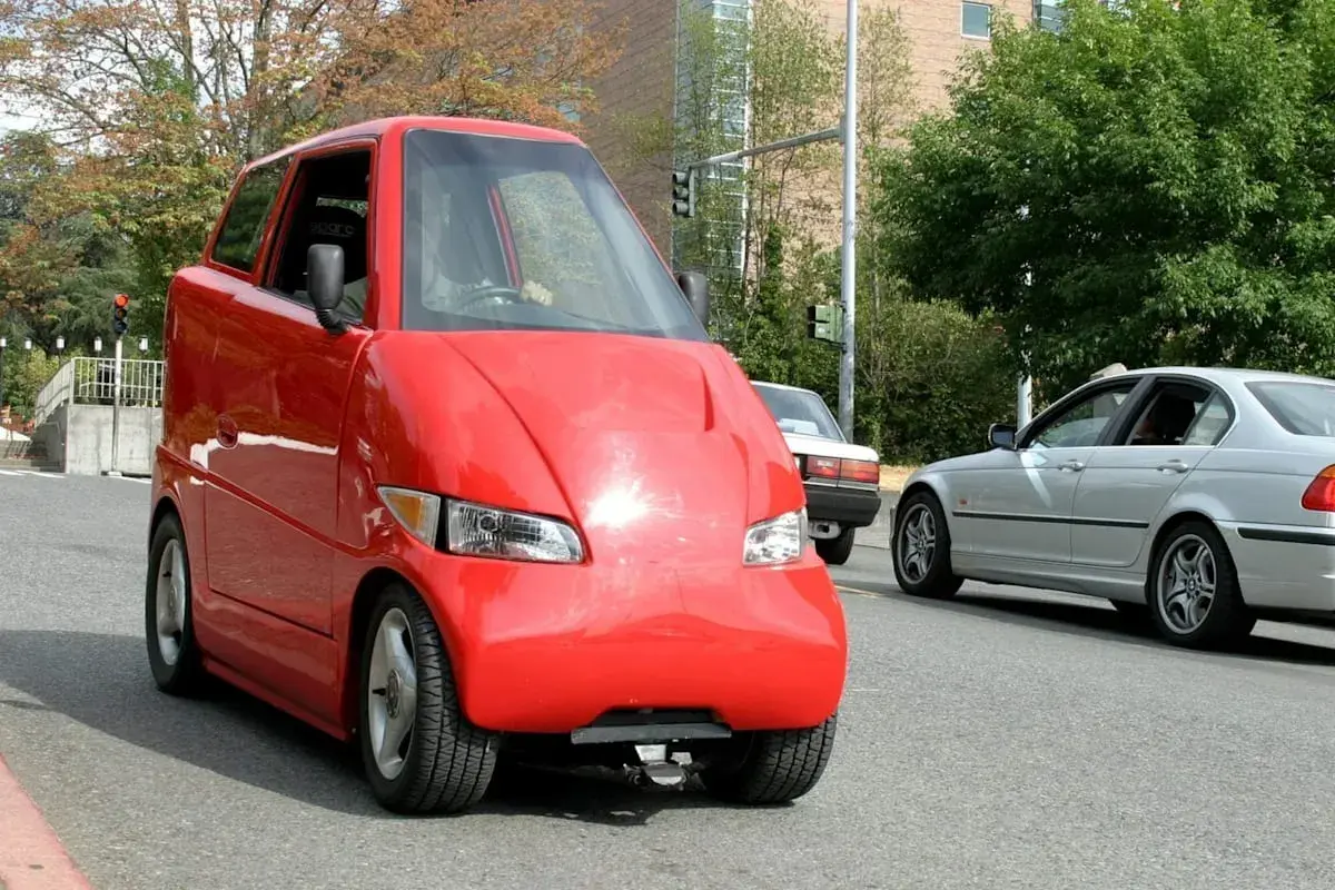 Roter, komischer Kleinwagen mit einer ungewöhnlichen Form fährt auf der Straße. Daneben ein silbernes Auto.