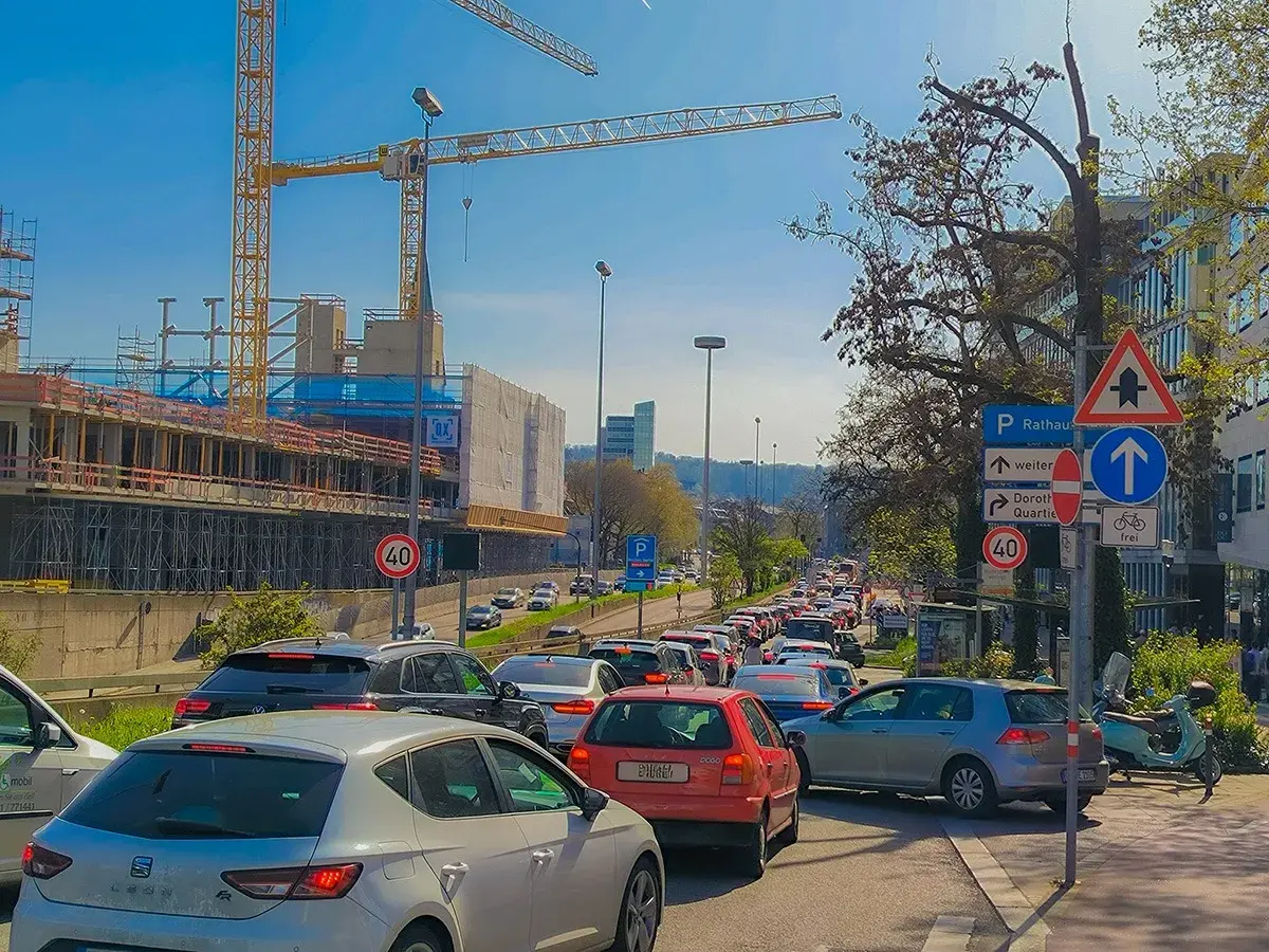 Stau auf einer mehrspurigen Straße mit Baustelle und Kränen im Hintergrund. Verkehrsschilder weisen auf Rathaus und Dorothy Quartier hin. Ein Roller parkt am Straßenrand.