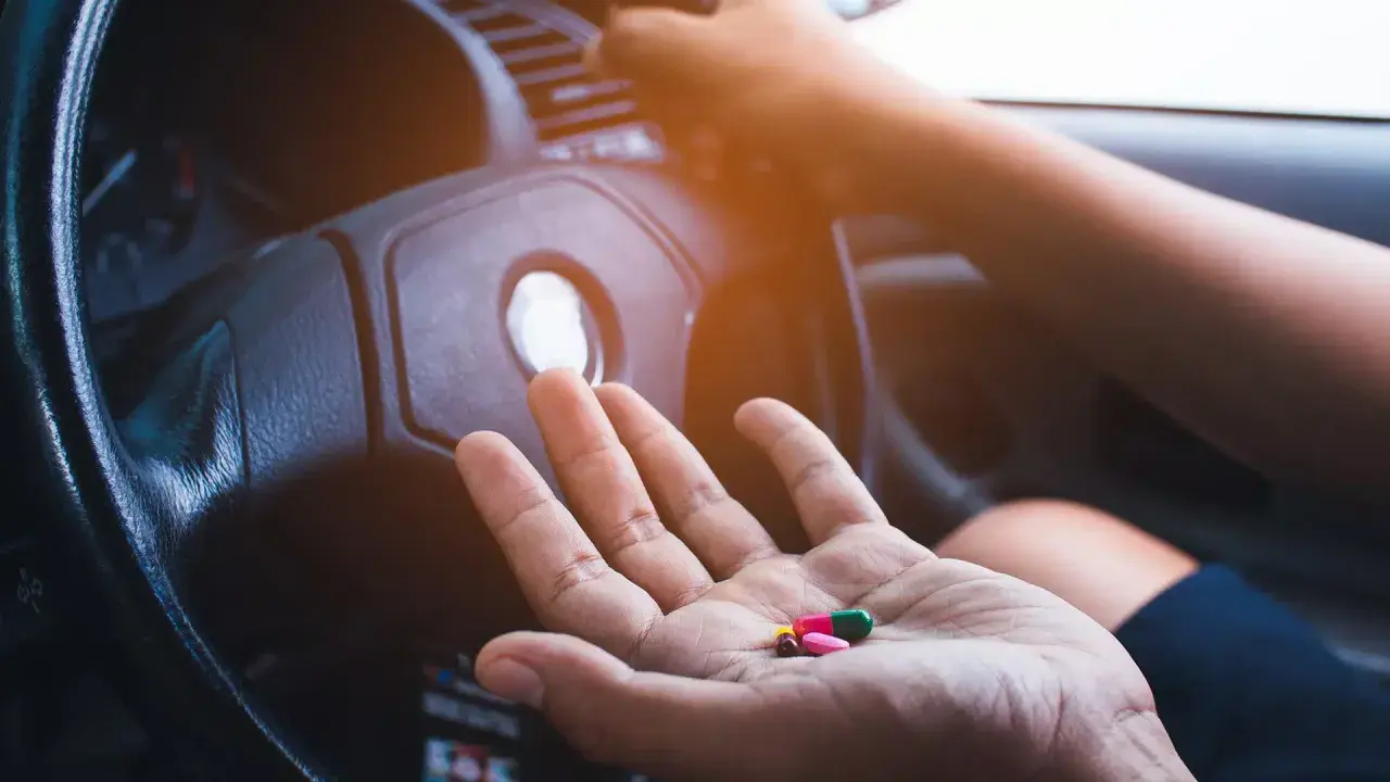 Hand hält Pillen, darunter Ibuprofen, vor einem Lenkrad. Vorsicht beim Autofahren mit Medikamenten!