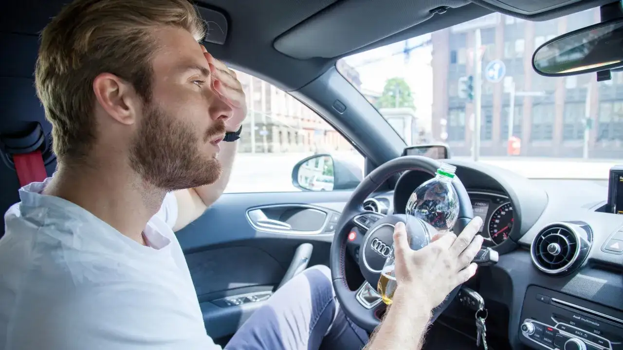 Mann mit Kopfschmerzen und Bierflasche am Steuer. Vorsicht bei Medikamenten wie Ibuprofen und Autofahren.
