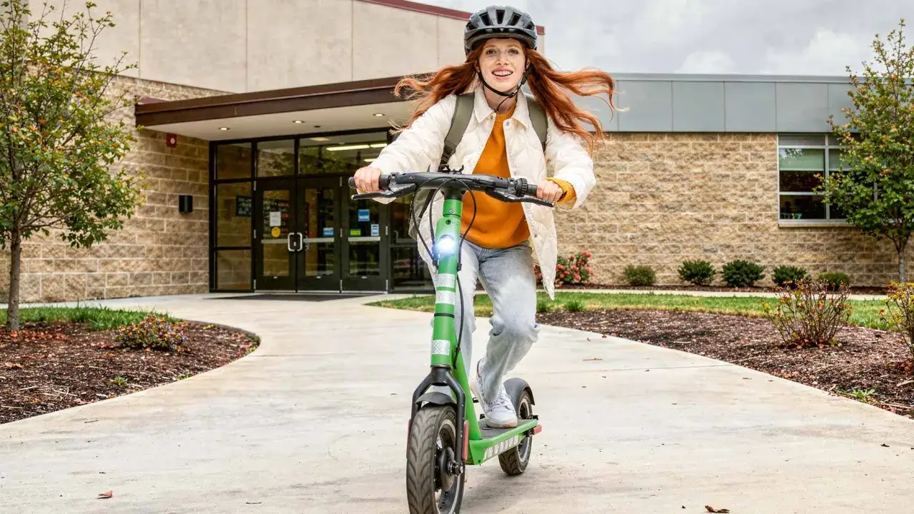 Ein Mädchen mit roten Haaren fährt auf einem grünen E-Scooter, bereit für die Schule. Sie trägt einen Helm und einen Rucksack.