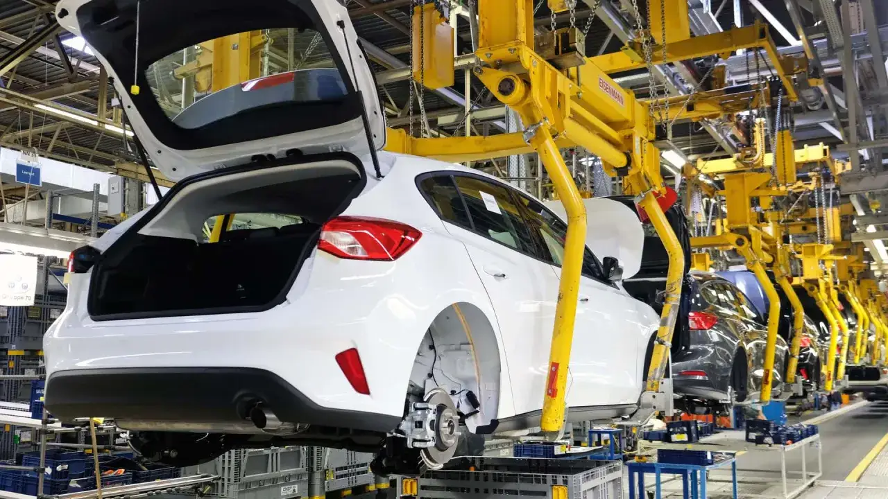 Ein weißer Ford Focus wird in der Fabrik montiert. Die Heckklappe ist geöffnet, und das Auto hängt an einer gelben Förderanlage.