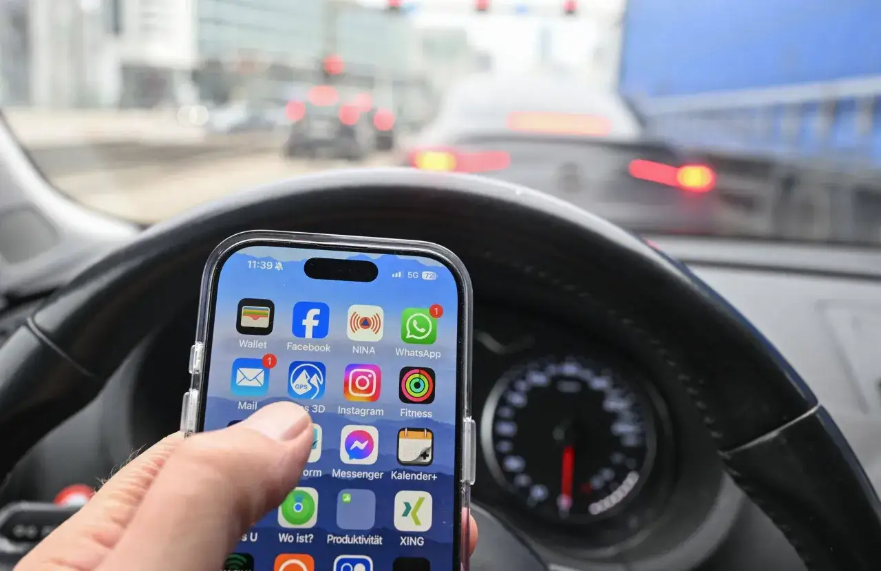 Hand h&auml;lt Smartphone mit Apps wie NINA und GPS, um einer Blitzer-Strafe zu entgehen. Blick auf das Lenkrad und die Stra&szlig;e.