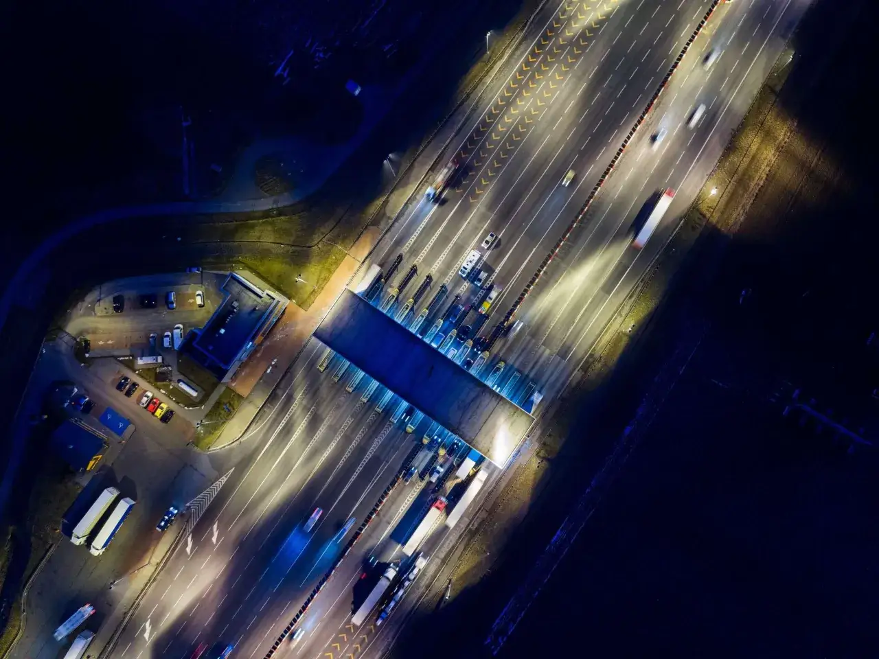 Nachtaufnahme einer Mautstelle in Belgien. Autos und Lastwagen passieren die beleuchteten Spuren, während die Lichter der Fahrzeuge lange Streifen auf der Straße hinterlassen.